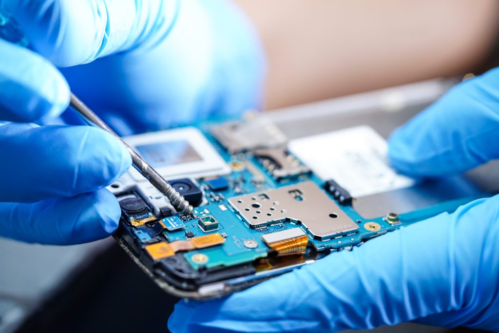 Person wearing blue gloves uses tweezers to adjust components on a smartphone circuit board.